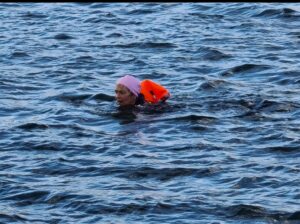 A blonde lady's head can be seen in an expanse of blue water. She has a pale pink swimming cap on and has an orange flotation device hear her head. 