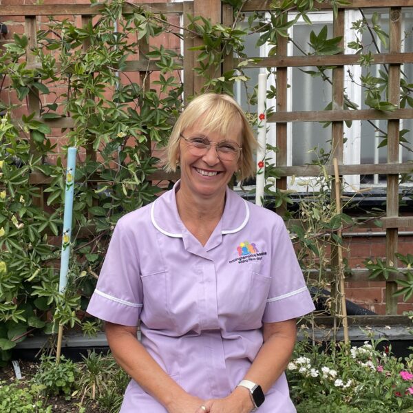 A lady with short blonde hair smiles at the camera - there are plants behind her. She wears a lilac nurses tabard.