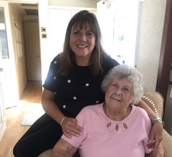 A lady with brown hair and blue top sits behind an older lady in a pink t-shirt. She has her arms round her shoulders.