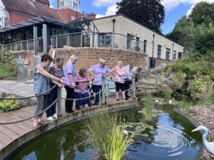 A group of people stand by the side of a large pond, with the Hospice building in the background. 