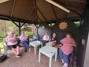 A group of patients and volunteers sit in a wooden summerhouse. There is a small green table and a yellow sun is placed on the wall. 