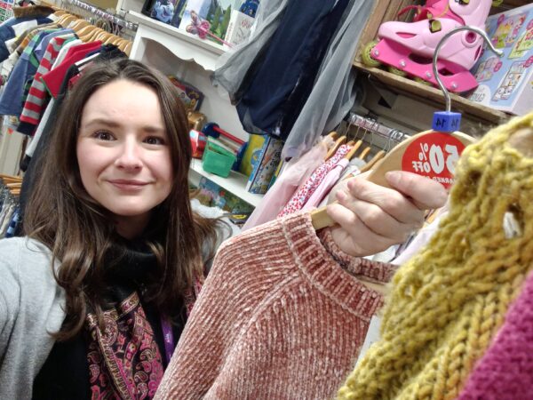 A woman with dark shoulder length hair, grey cardigan and black scarf hangs up a pink chenille jumper, with a 50% off sticker.