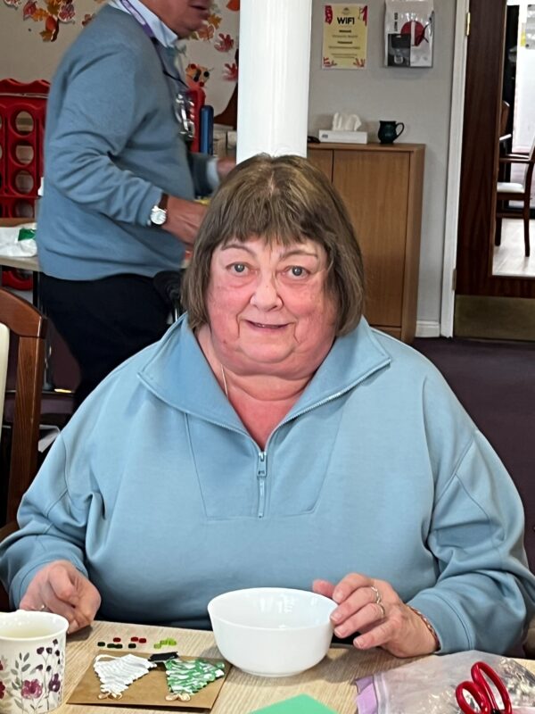 June has short brown hair and a fringe, and in this picture wears a blue coat with craft supplies on a table in front of her.